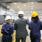 Male engineer and female factory employees in hardhats walking on plant floor and talking, man pointing at equipment and instructing women. Back view. Industrial occupation concept